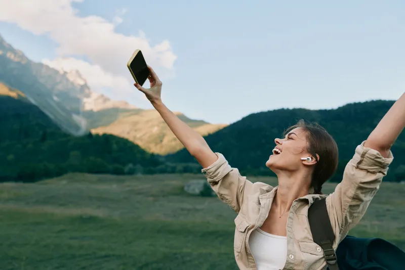 Joyful woman outdoors, taking a selfie with a phone, in a sunny meadow with a mountain backdrop, relaxed posture, natural colors, fresh breeze.