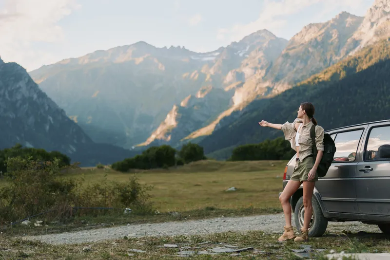 Young woman with a backpack stands next to a car on a mountain road while pointing towards the distant peaks during a clear day.