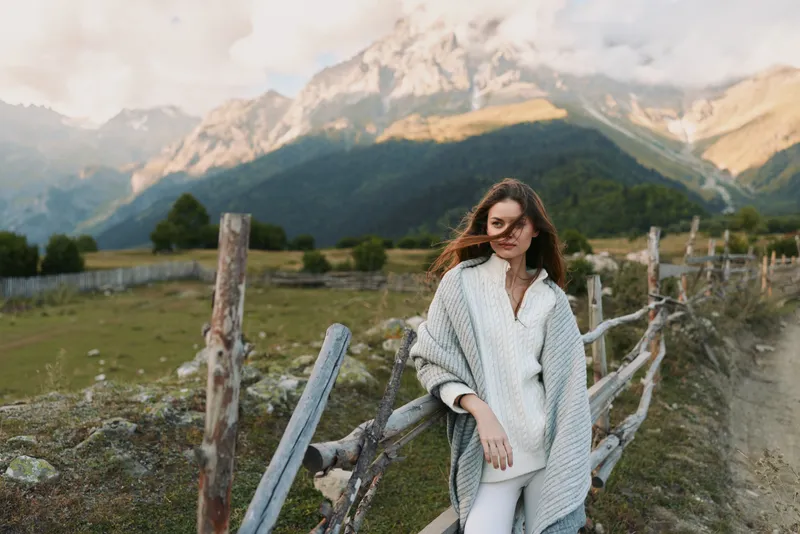 Cheerful woman outdoors, wearing a light cardigan by a wooden fence, mountains in the background, soft breeze, relaxed lifestyle moment, natural colors