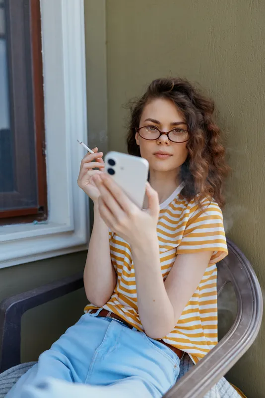 Young woman with curly hair and glasses sits on a balcony, looking at her smartphone while holding a cigarette, relaxed pose, soft smile, casual striped top.