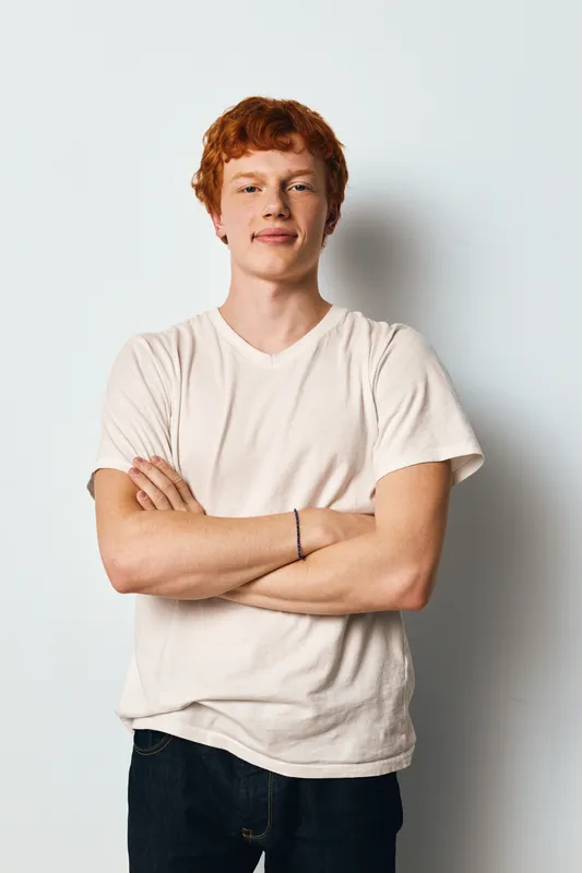 Young man with red hair standing with arms crossed wearing white t shirt and jeans against white background looking confident and relaxed in casual style portrait.