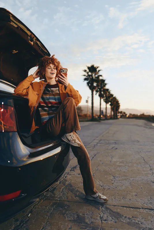 Woman lifestyle scene near open car hood, taking a phone selfie on a quiet road at sunset, light and palm trees in background, casual fashion