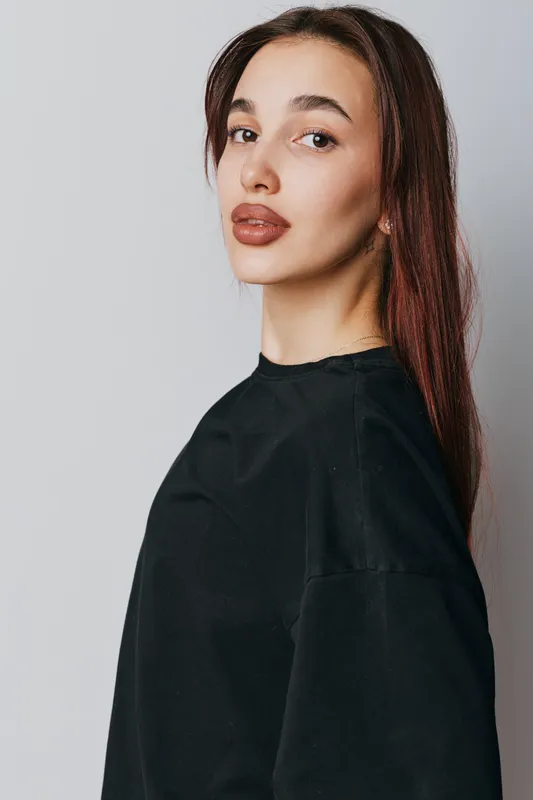 Young woman with long brown hair wearing black sweater stands against gray background looking at camera with neutral expression. Portrait of female model in casual clothing with natural makeup.