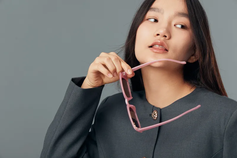 Young woman with long dark hair holding pink glasses wearing gray jacket standing against gray background looking sideways. Portrait of thoughtful female with casual elegant style fashion.