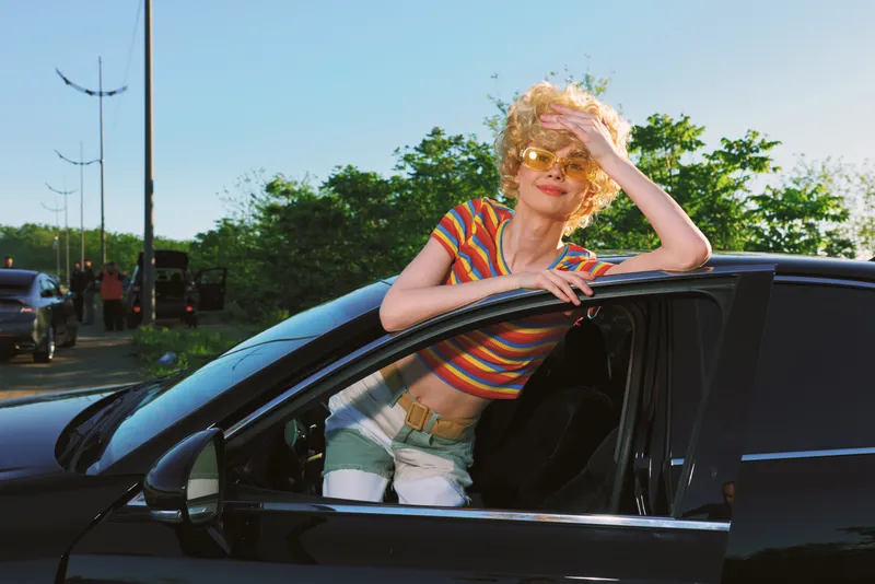 Smiling woman leaning out of a black car window in a striped top, enjoying sunshine. Outdoor street scene with retro color and casual fashion, carefree mood!