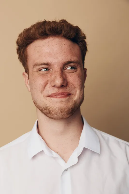 Young man with red hair and beard wearing white shirt looks sideways with slight smile on beige background. Concept of casual style, natural expression, and male portrait photography.