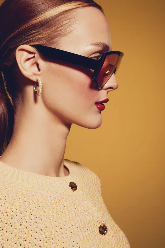 Profile of a stylish woman wearing large sunglasses and gold hoop earrings against a warm yellow background. She wears a textured sweater with buttons, showcasing fashion and elegance.