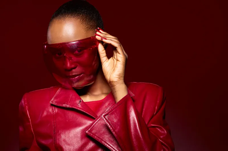 Portrait of a dark skinned African woman wearing a red leather jacket and red translucent visor, posing confidently on a red background. Fashion concept with bold color and style.