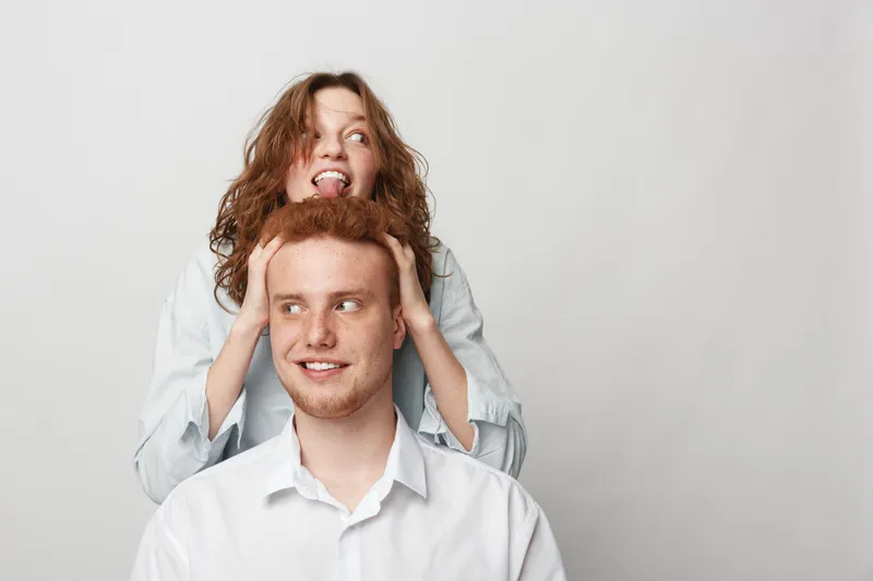 Happy young redhead man and woman with curly hair playfully interact in white shirts on light background. Concept of friendship, fun, casual lifestyle, and positive emotions.