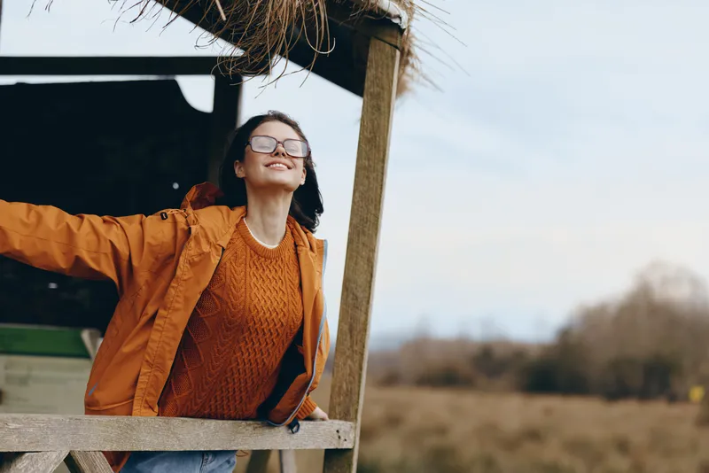 Woman lifestyle portrait outdoors, smiling in orange sweater and glasses, leaning from wooden shelter, joyful mood, autumn field background, natural light, rural walk, calm, fresh air today