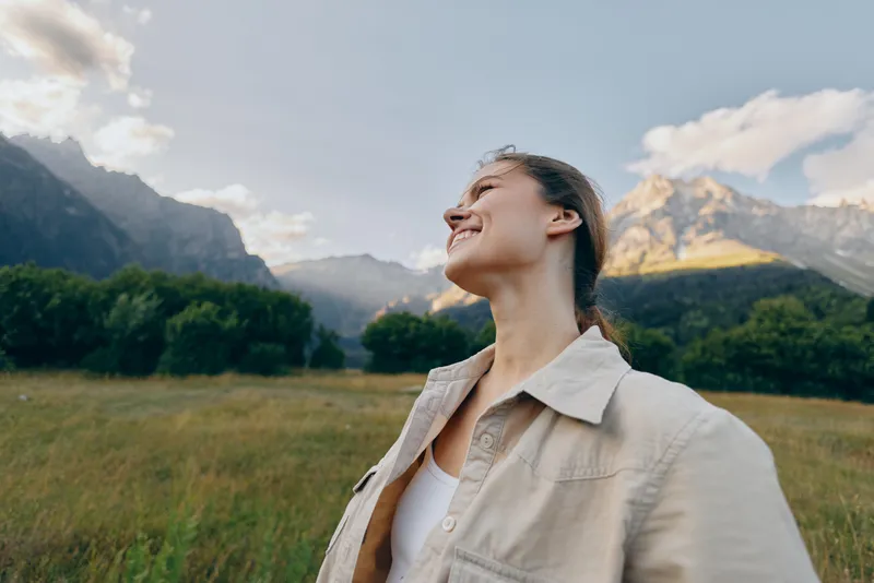 Young woman with brunette hair enjoys fresh air and sunshine while standing in a green meadow with mountains in the background, expressing happiness and freedom.