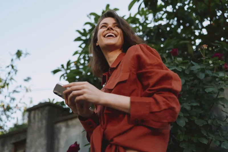 Cheerful woman outdoors in a red coat, smiling with eyes bright, holding a smartphone, sunlit moment, casual lifestyle, natural colors, lush greenery background soft breeze