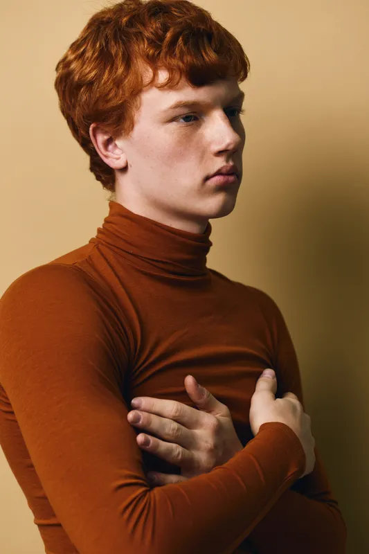 Portrait of young redhead man in brown turtleneck sweater with arms crossed on chest looking aside on beige background. Indoor studio shot with soft light on face and thoughtful expression.