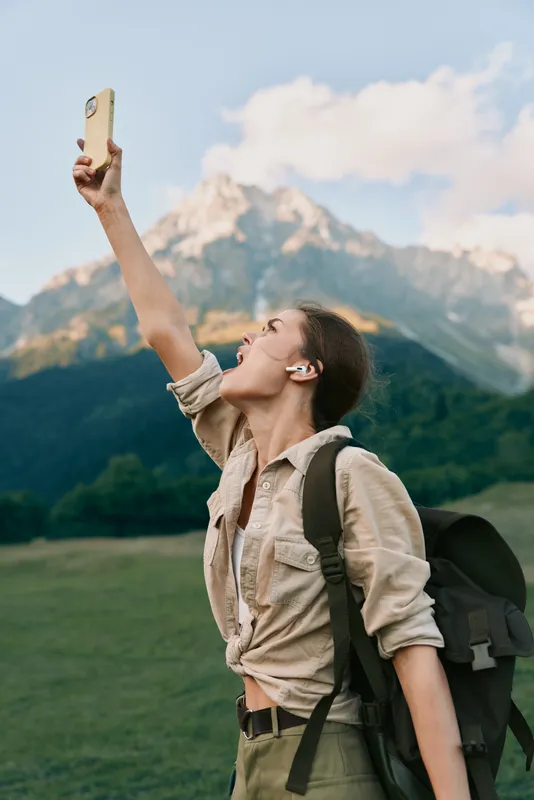 A young woman with a backpack takes a selfie in a grassy meadow as rugged mountains loom in the background, capturing a moment of adventure and confidence.
