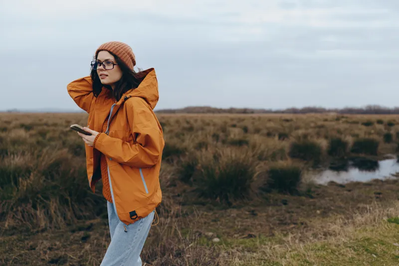 Woman lifestyle outdoors in autumn field, orange jacket and hat, standing by a river path, hands in pockets, relaxed mood in countryside nature.