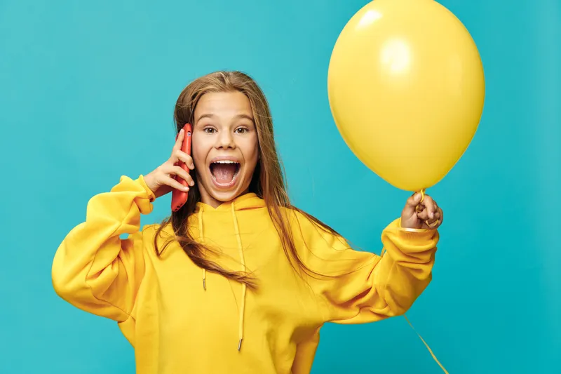 Girl in yellow hoodie holding balloon and smartphone, smiling with excitement against blue background for celebration, communication and happiness concept