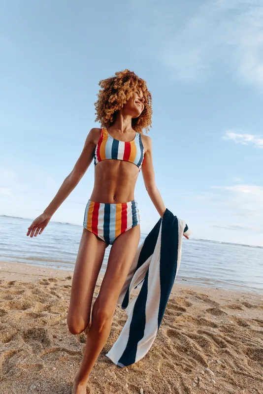 Joyful woman on a sunny beach wearing a colorful striped bikini, running with a towel, wind in her curls, clear sky, blue ocean in background, energetic summer moment