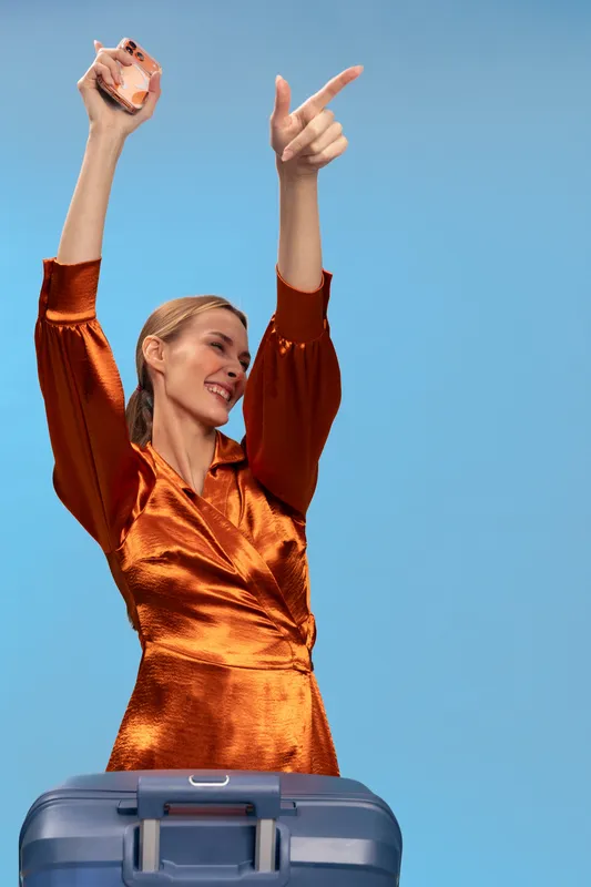 Happy woman in orange satin dress holding perfume bottle and raising arm, standing behind blue suitcase on light blue background. Concept of travel, fashion, and joyful mood.