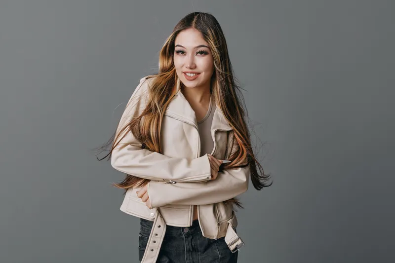 Young woman posing with long hair and beige jacket on grey background smiling confidently with casual style for fashion portrait and lifestyle photography studio.