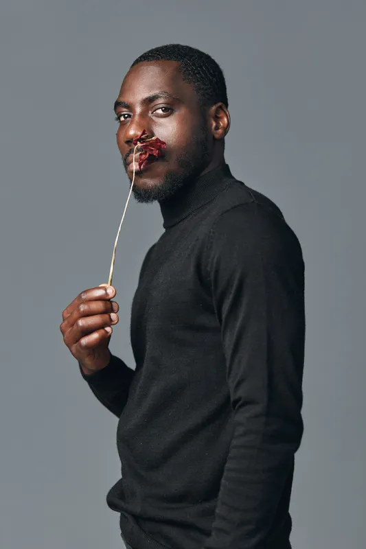 African man with dark skin wearing black turtleneck holding dried flower on stick near face in creative conceptual visual style studio portrait with gray background.