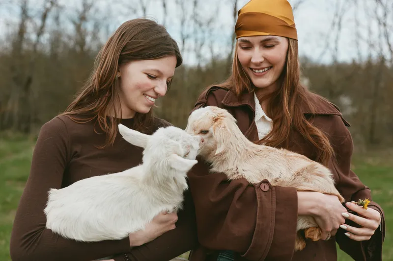 Two women smile as they cradle friendly goats in a sunny countryside scene, showcasing rural lifestyle and bonds between people and animals, tender moment of care and farm life