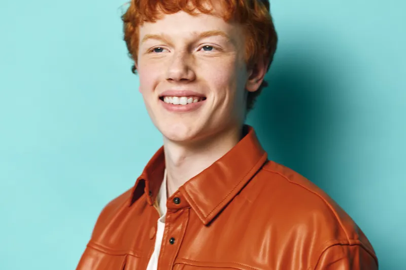 Young man with red hair smiling and wearing orange leather jacket against blue background. Casual portrait of happy person with natural expression and clear skin in studio lighting.