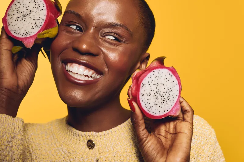 Happy dark skinned African woman holding two halves of dragon fruit near face smiling with white teeth on yellow background. Concept of healthy food, tropical fruit and positive emotion.