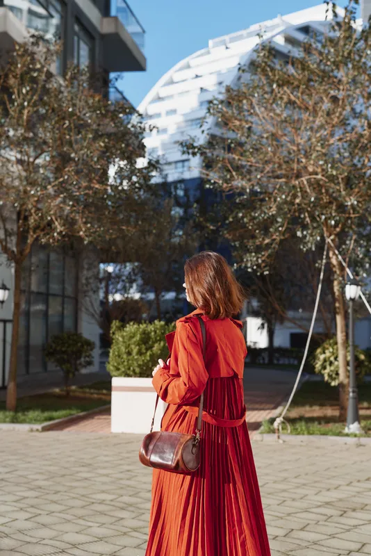 Young woman with red hair wears a long red dress and carries a brown leather bag while walking outdoors in a sunny urban park with autumn trees around her.