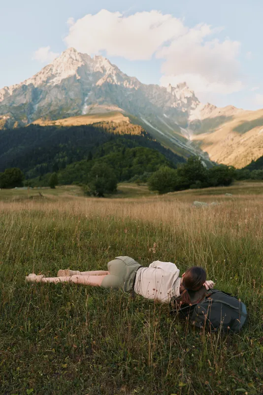 Calm woman lying in a sunlit meadow with distant mountains, relaxed pose, gentle breeze, natural colors, lush grass, outdoor lifestyle moment soft light, open landscape.