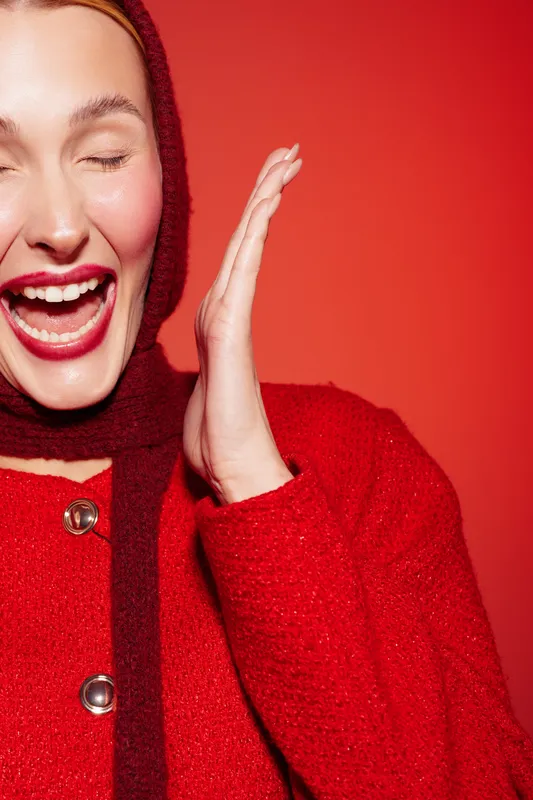 Happy woman in red sweater and scarf laughs with closed eyes on red background. Close-up portrait with joyful expression and vibrant color. Concept of happiness and winter fashion.