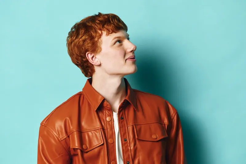 Young man with red hair in brown leather jacket looking up against blue wall, casual portrait with side profile and soft lighting for style and personality concept.