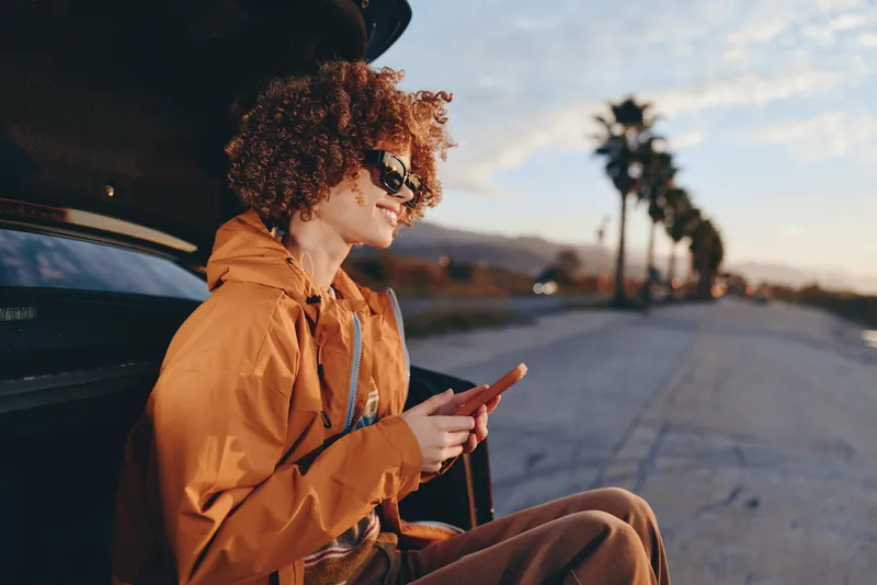 Woman lifestyle scene outdoors, sitting beside a car on a road at sunset, wearing an orange jacket, holding a smartphone, curly hair and profile visible, calm travel moment