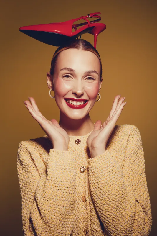 Smiling woman with red high heel shoe on head poses with hands near face against yellow background. Concept for fashion, fun, creativity, and joyful expression in studio lighting.