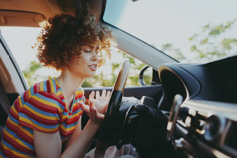 Woman driver with curly hair sits in car interior, wearing striped shirt, candid lifestyle shot with retro film color, warm sunlight, genuine smile, dynamic pose, urban drive vibe