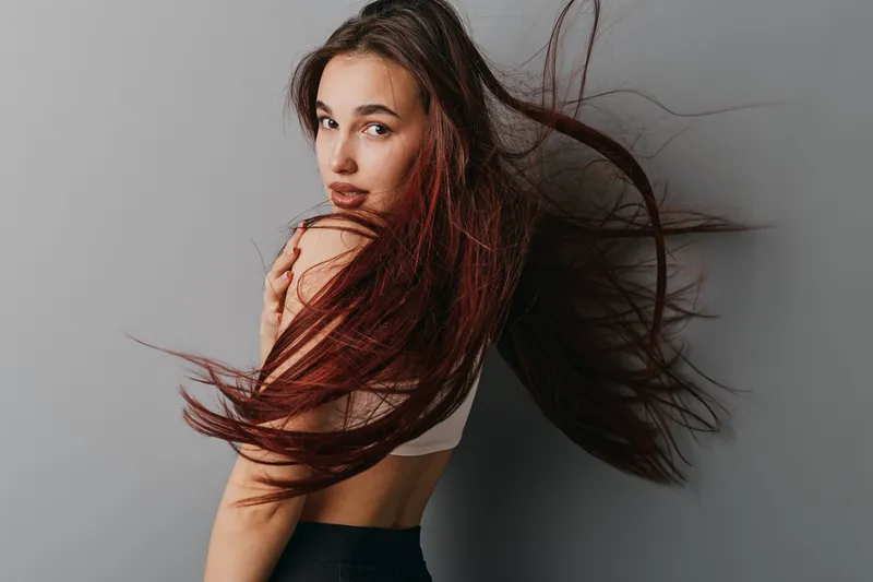 Young woman with long hair blowing in wind posing on gray background. Beauty portrait with natural makeup and confident expression in casual sleeveless top and dark pants.