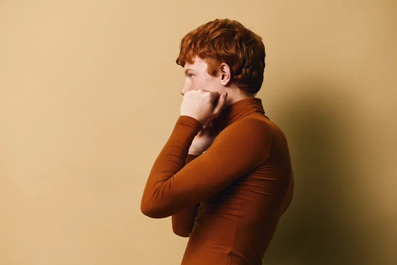 Profile of redhead woman with short curly hair wearing brown turtleneck, standing with closed fists near face against beige background indoor.