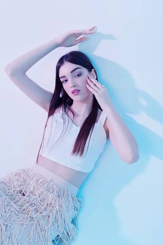 Young woman with long dark hair poses in white crop top and textured skirt under soft blue lighting. Concept of beauty, fashion, and modern style photography.