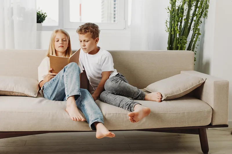 Two children sit barefoot on beige sofa in bright living room, reading book together. Casual clothes, natural light, cozy home atmosphere, childhood learning and sibling bonding concept.