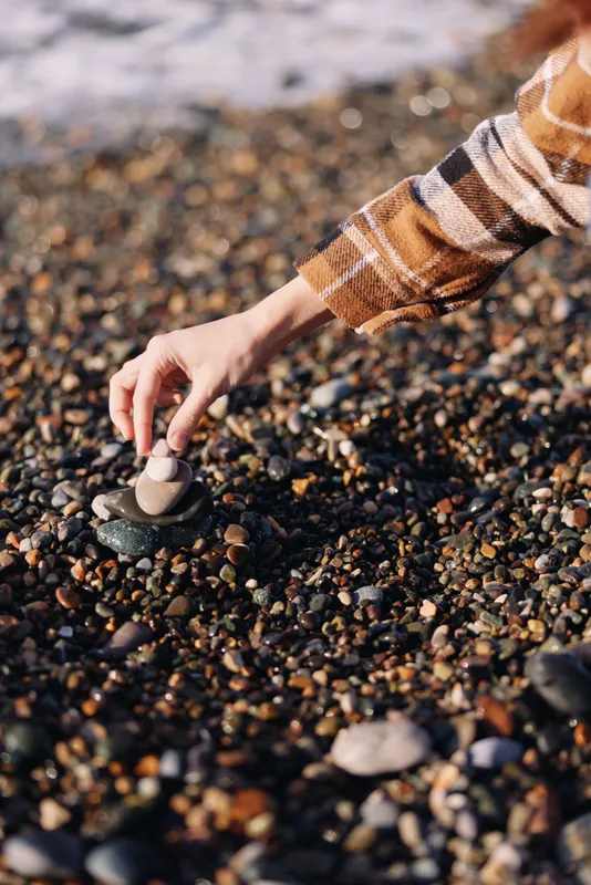 Close view of a hand placing a small stone on a pebbled beach with gentle waves, captured in warm light, highlighting patience, mindfulness and a simple coastal ritual to evoke calm and connection.
