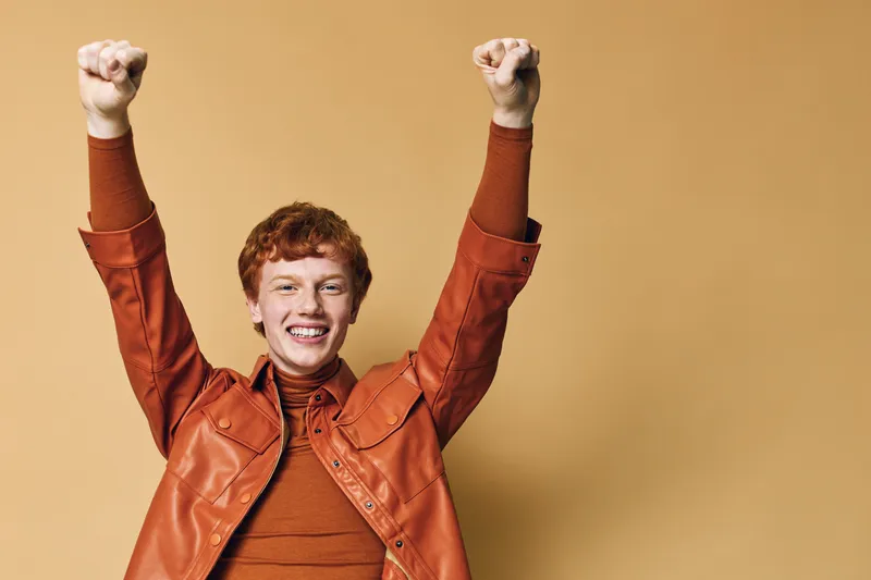 Happy man with red hair in brown jacket raising fists in celebration against beige background with excitement, joy, success, and enthusiastic expression in casual style.