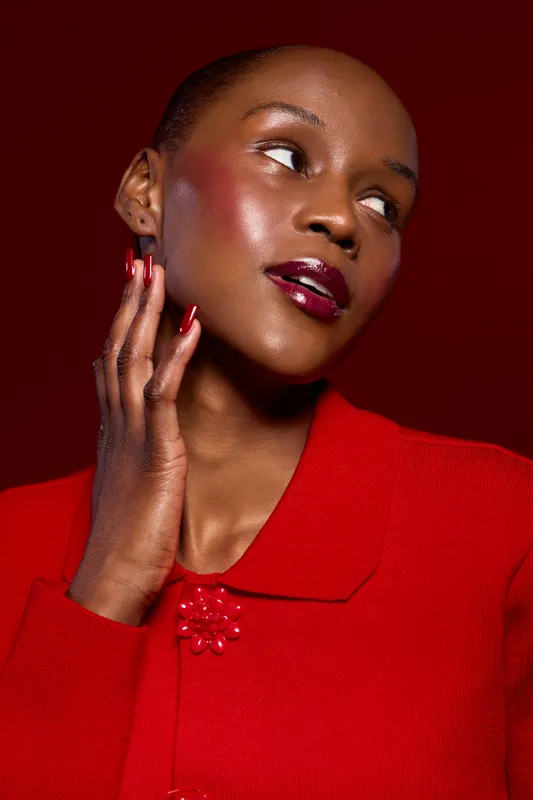 Close-up portrait of a dark skinned African woman with glossy lips and red nails wearing a red sweater against a red background. Concept of beauty, fashion, and elegance with dark skin tone.