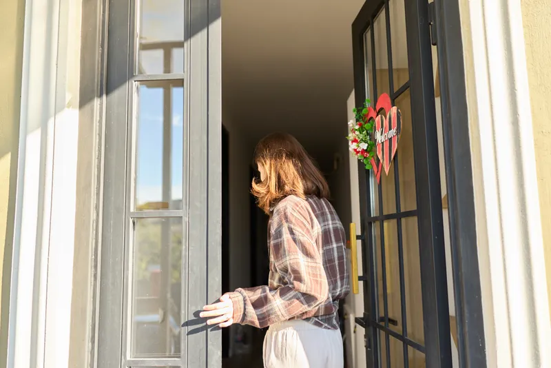 Candid woman entering a doorway outdoors, casual lifestyle moment, sunlight on plaid shirt, soft breeze, inviting home entry, natural colors, warm atmosphere, everyday charm