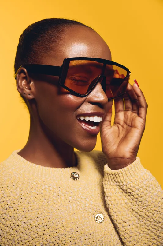 Happy African woman with dark skin wearing oversized sunglasses and cozy knitted sweater smiles with closed eyes on yellow background. Concept of fashion, joy, and beauty.