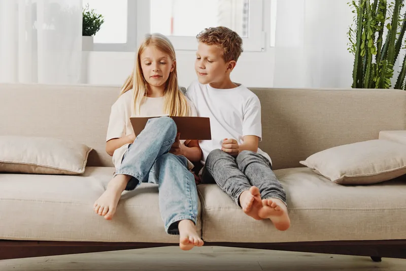 Two children sitting barefoot on beige sofa reading a tablet together in bright living room with plants and cushions around.