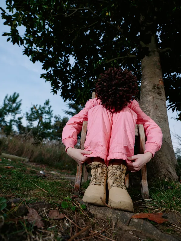 A person sits on a wooden chair outdoors, wearing a pink shirt and rugged boots, with curly hair and a relaxed pose, surrounded by trees and green grass in a peaceful natural setting.
