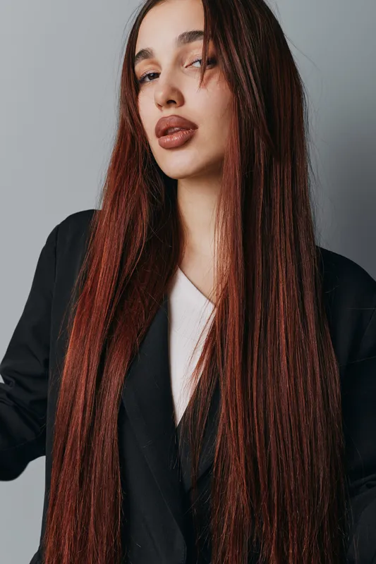 Woman with long straight hair and natural makeup posing in black jacket against gray background. Closeup portrait of confident female with brown hair and neutral expression.