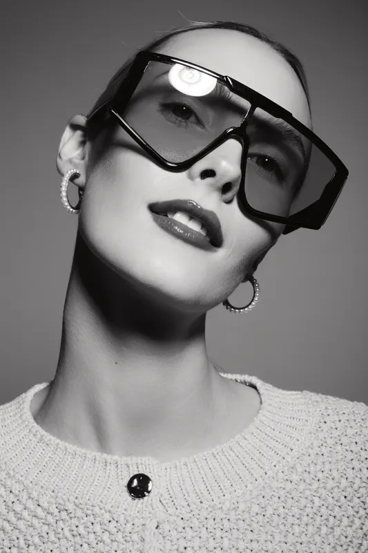 Young white woman with pale skin wearing large sunglasses and hoop earrings poses in studio. Fashion, style, and beauty concept in black and white photo.