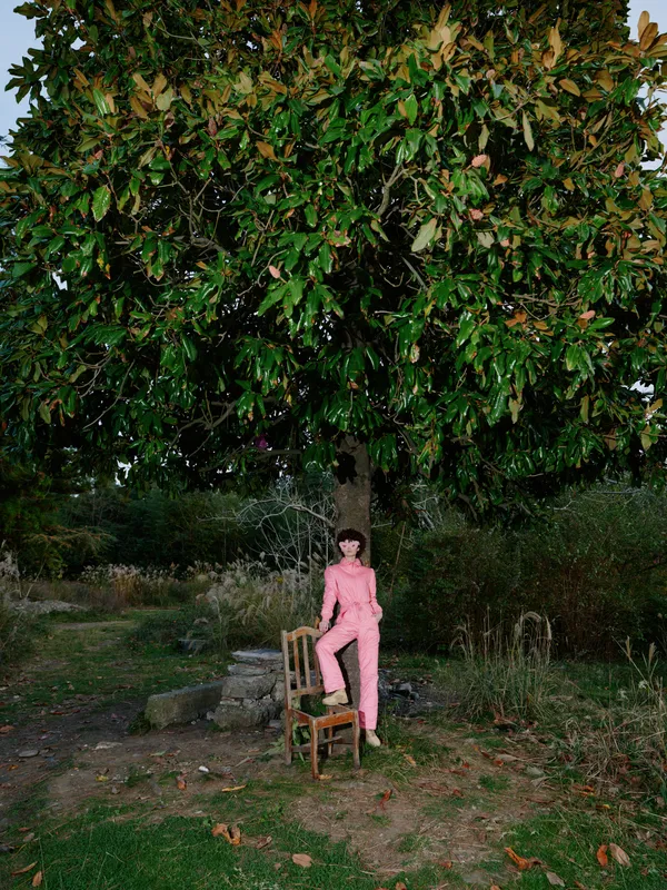 Woman in pink outfit seated on a chair beneath a large leafy tree in a garden, shaded by branches, creating a peaceful outdoor portrait with natural light and calm atmosphere. Soft breeze.