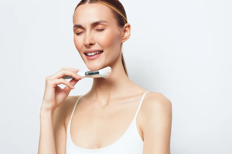 A smiling woman with closed eyes applies cosmetic cream on her face using a brush. She wears a white tank top against a plain white background. Concept for skincare and beauty routine.
