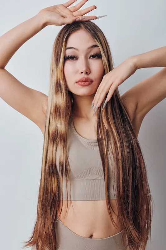Young woman with long straight hair posing with hands near face and head in beige sleeveless top against neutral background for fashion and beauty.